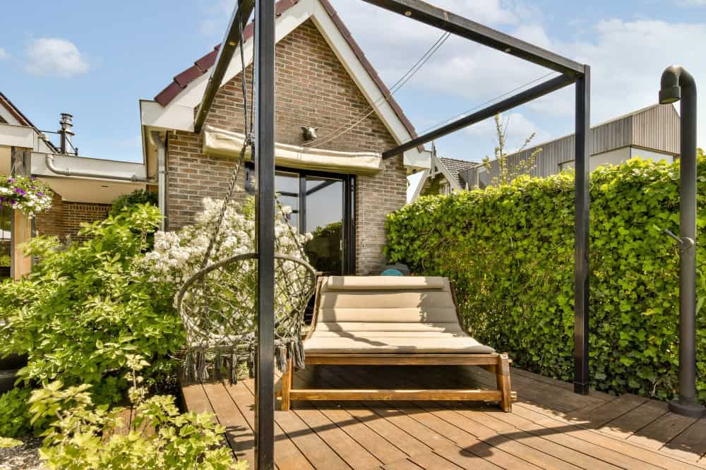 Een gezellige patio met een houten dek is voorzien van een tweezits buitenschommel onder een zwart metalen frame. Weelderige groene hagen en bloeiende planten omringen het gebied en een bakstenen huis met grote glazen deuren staat op de achtergrond onder een blauwe hemel.