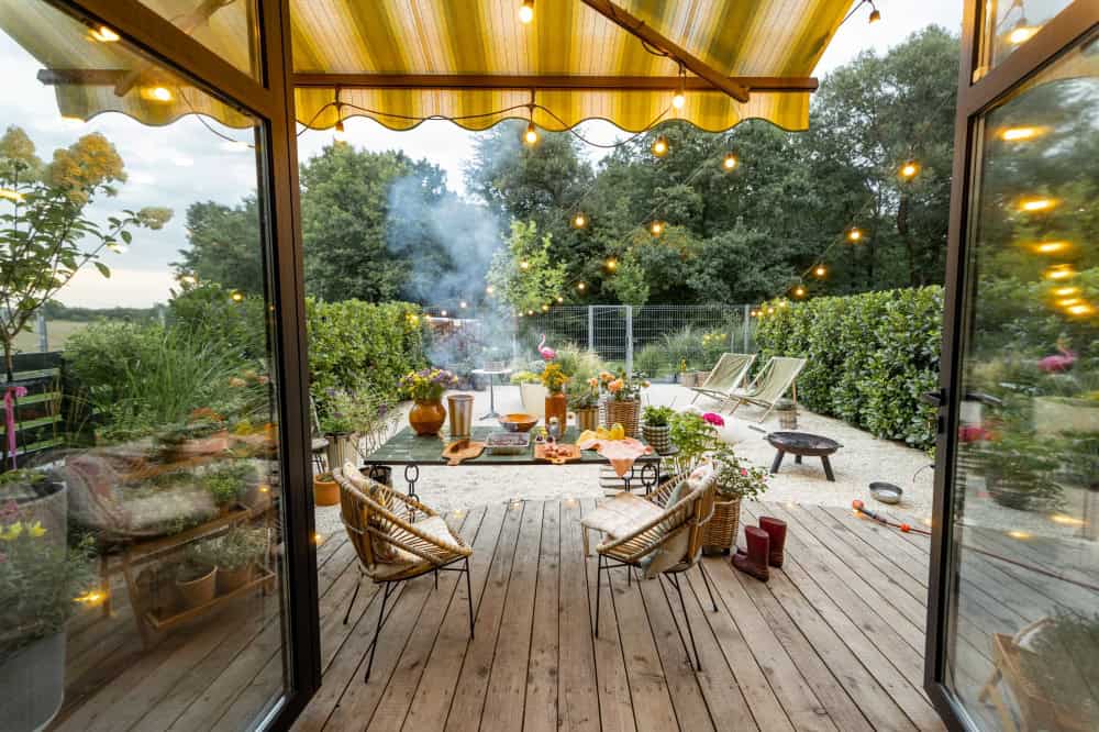Een gezellig terras met lichtjes, rieten stoelen, planten in potten en een houten tafel gedekt voor een maaltijd. Op de achtergrond stijgt rook op van een grill. Een zonnescherm biedt schaduw en weelderig groen omringt de buitenruimte. De ruimte ziet er uitnodigend en feestelijk uit.