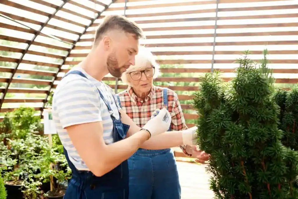 Een jonge man in overall en handschoenen onderzoekt een plant terwijl een oudere vrouw met bril en geruit overhemd aandachtig toekijkt. Ze bevinden zich in een zonovergoten, houten kas met verschillende potplanten op de achtergrond.