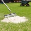 Een dunne laag zand wordt met een gazonrakel over een lage plek in het gras uitgetrokken, kruiwagen op de achtergrond