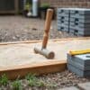 Handstamper op een zandbed tussen houten kantplanken in een kleine achtertuin, met een stapel betontegels ernaast