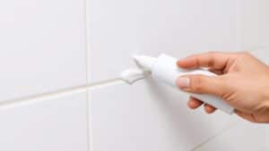 Hand die witte kit aanbrengt in de hoek van een douche, met een verse naad tegen de tegels.