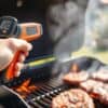 Hand met infraroodthermometer boven een gloeiende barbecue met steaks in een Nederlandse tuin.