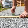 Persoon verspreidt 5 cm grind op een tuinpad met een waterpas, met zakken grind op de achtergrond.