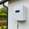 Thuisbatterij aan de muur van een Nederlands huis met display, in een tuin zonder zonnepanelen.